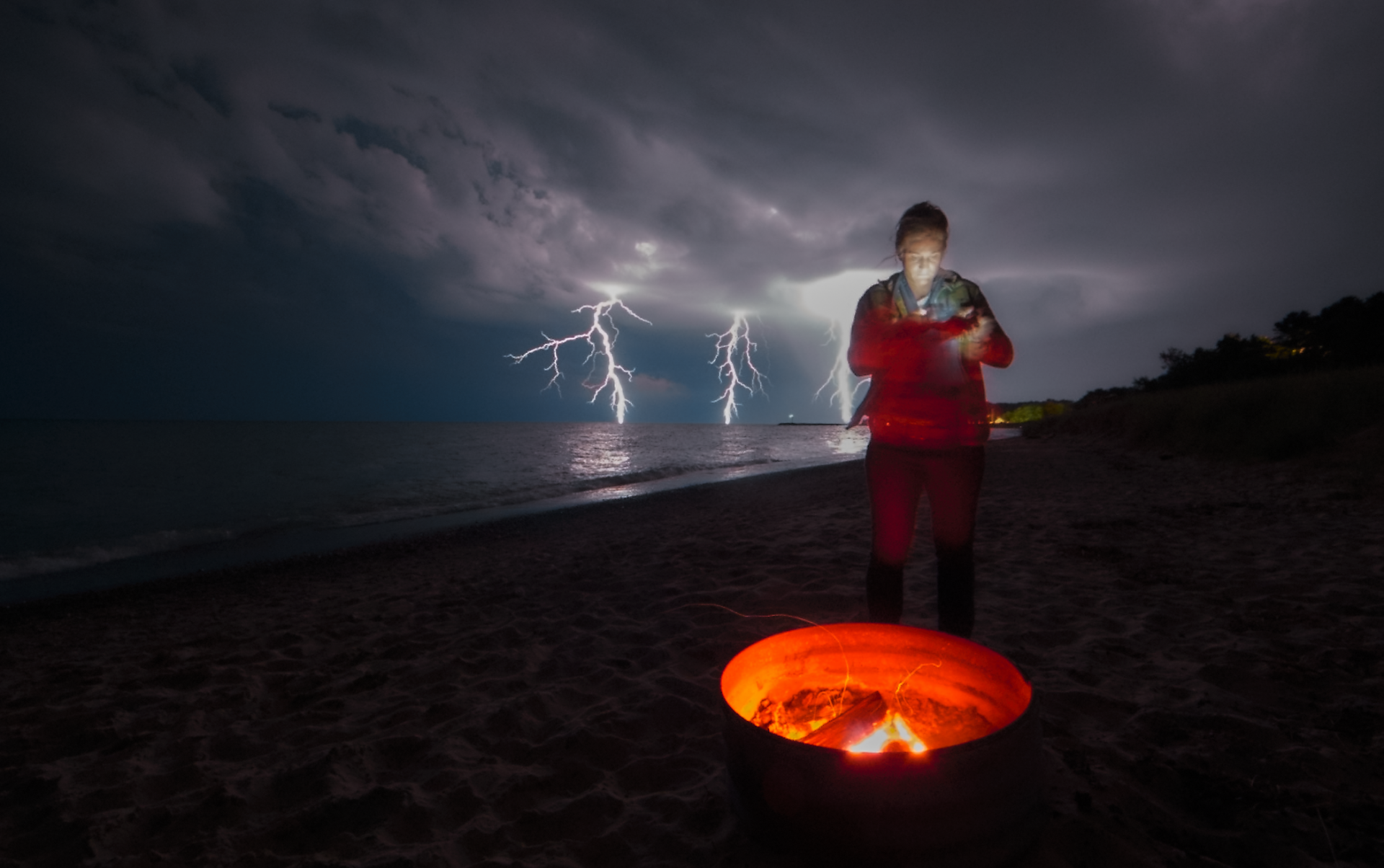 lightning at night with woman texting in front of black fire at vans beach in Leland Michigan